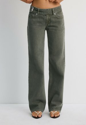 Personne portant un jean vert taille haute à jambes larges et des sandales blanches à brides, debout devant un fond clair uni.