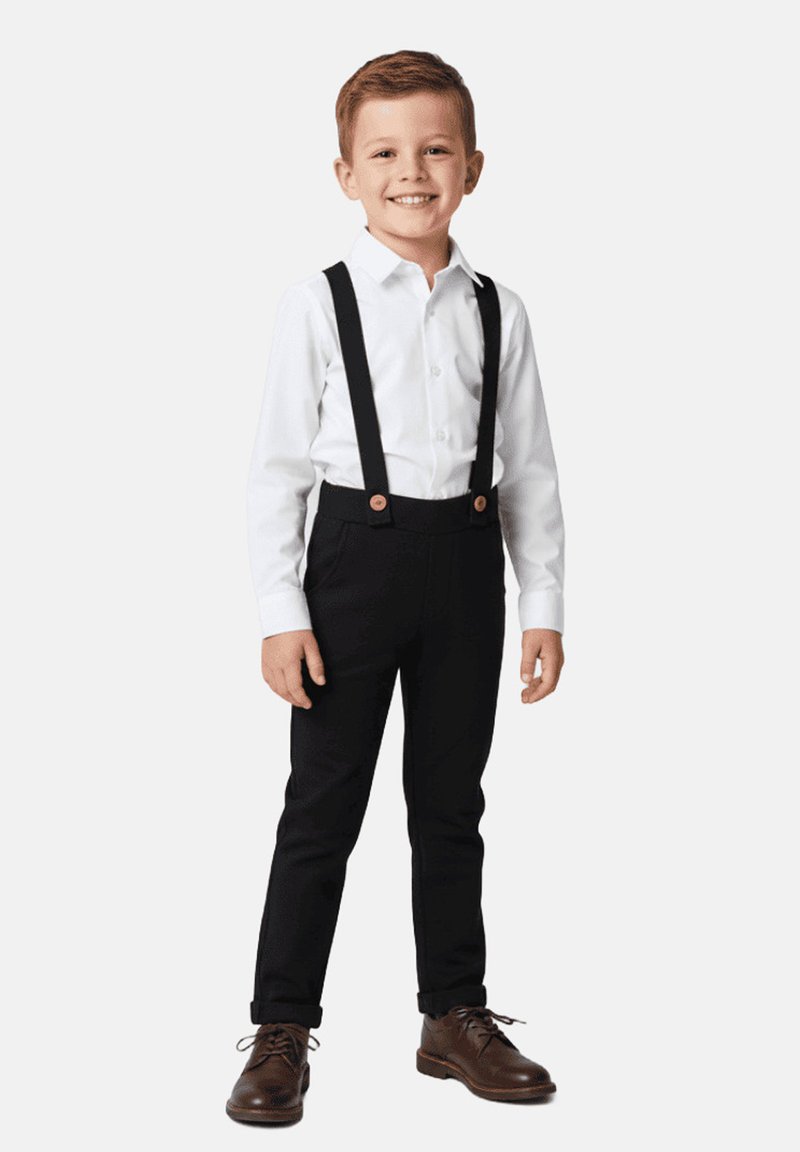 Biała koszula na guziki, czarne spodnie na szelkach, brązowe buty, brązowe guziki na szelkach. Uśmiechnięte młode dziecko.