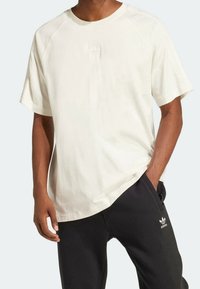 Man i en lös vit Adidas t-shirt och svarta Adidas joggingbyxor med logotyper på båda plagg, står med en hand i fickan.