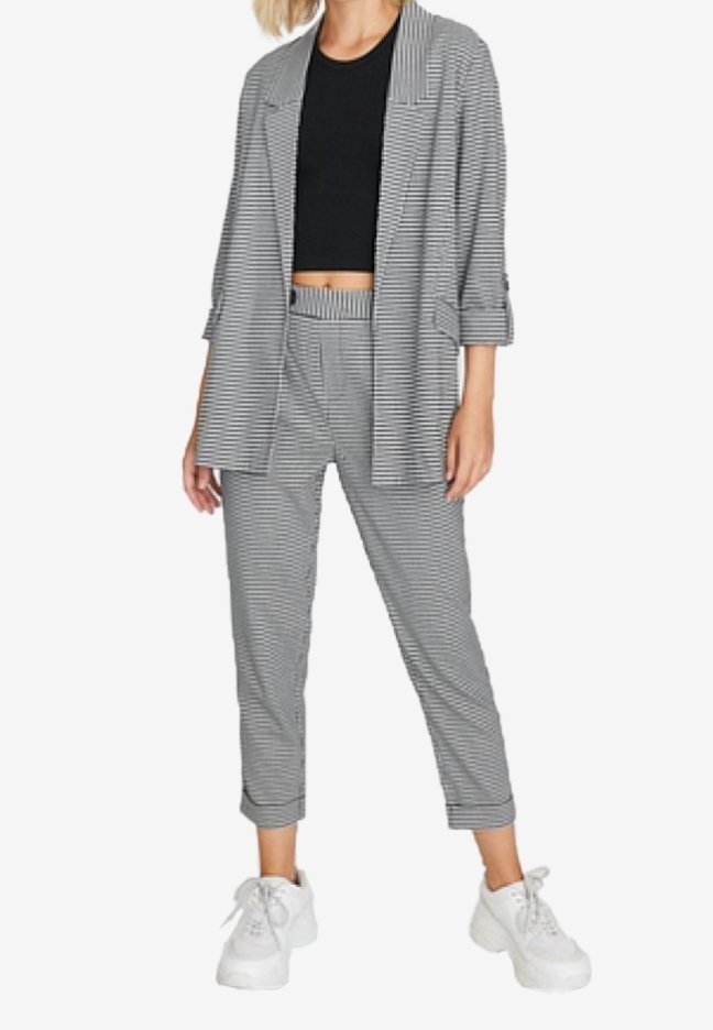 Personne portant un blazer rayé gris et blanc et un pantalon, un crop top noir et des baskets blanches épaisses, debout devant un fond blanc.