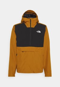 Veste d'extérieur à capuche jaune moutarde avec un panneau noir sur la poitrine et le logo blanc The North Face en haut à droite.