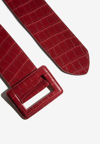 Curea lată roșie cu textură în model de crocodil și cataramă mare rectangulară așezată pe o suprafață albă.