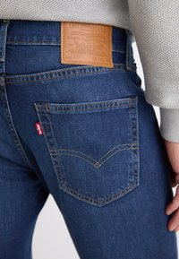 Blå denimbukser med en lærreim med teksten "Levi Strauss & Co." bak. Har en buet sydd lomme og gyllent søm.