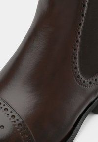 Boots de gleznă din piele maro, cu o textură netedă, detalii perforate, vârf rotund și panouri elastice pe laturi pentru o purtare ușoară.
