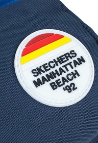 Tela azul marina con tejido texturizado que presenta un parche blanco circular. El parche incluye el texto negro "SKECHERS MANHATTAN BEACH '92" y franjas de colores.