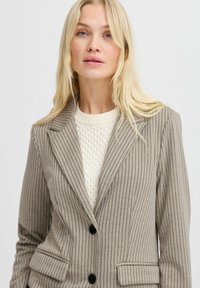 Kohandatud hall bleiser vertikaalsete pinstripe'idega, klanitud krae, kaks esikäe taskut ja mustad nupud, kombineeritud kreemja tekstureeritud svetriga.