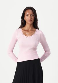 Rosa ribbad långärmad topp med V-ringning och spetsdetalj, i kombination med en svart plisserad kjol som framhäver en figurnära design och texturkontrast.