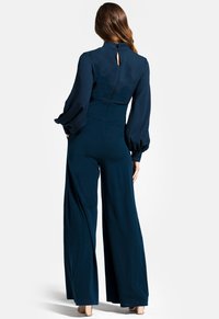 Navyblauer Jumpsuit mit langen, gepufften Ärmeln, hohem Kragen und Schlüsselloch-Detail am Rücken; weit geschnittenes Design aus weichem, strukturiertem Stoff.