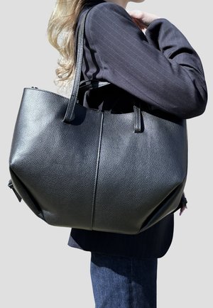 Persona bionda che indossa un blazer nero e porta una grande borsa tote in pelle nera sulla spalla contro uno sfondo semplice.