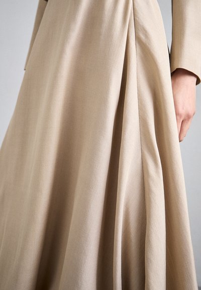 Robe beige plissée avec une texture lisse. Présente un tissu doux et fluide qui tombe élégamment, avec un gros plan sur les plis et une main la tenant.