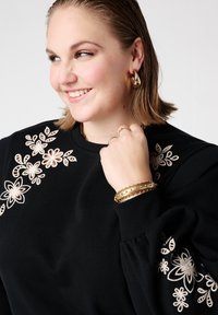 Zwarte sweatshirt met geborduurde bloemen in crèmekleur en gouden accenten. Model draagt gouden armbanden en oorbellen. Geverfd, zachte stof met ballonmouwen.