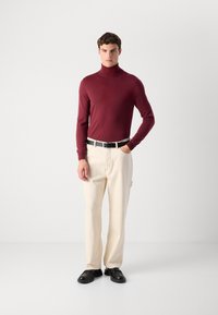 Mann som står med hånden i lommen, iført en bordeaux turtleneck genser, beige bukser, svart belte og svarte sko mot en enkel bakgrunn.