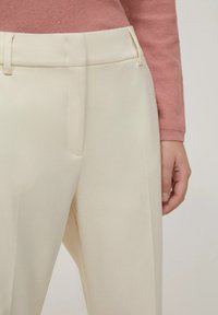 Pantaloni di un colore crema chiaro con una texture liscia, dotati di una vestibilità piatta, tasche laterali e passanti per la cintura. Mano posizionata sull'anca.