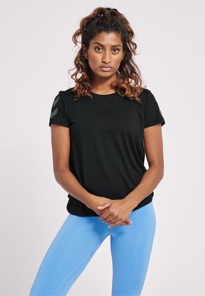 Kvinde med krøllet hår står med hænderne foldede, iført en sort T-shirt og lyseblå tights mod en ensfarvet lys baggrund.