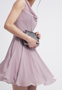 Světle růžové šatům bez rukávů s výstřihem do cowl; splývavá tkanina. Drží černou a stříbrnou kabelku s hadím vzorem.