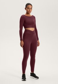 Set sportiv bordo format dintr-un top crop cu mânecă lungă și decolteu larg, împreună cu colanți cu talie înaltă și design încrucișat al taliei.