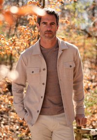 Man in een beige jack en een bruine shirt staat buiten tussen de herfstbladeren, met zijn handen in zijn zakken, kijkend naar de camera.