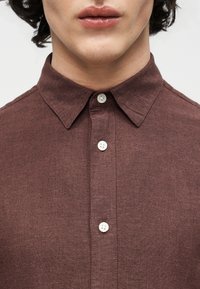 Gros plan sur la partie inférieure du visage et le cou d'une personne portant une chemise marron à boutons blancs et col pointu.