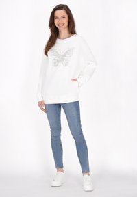 Felpa bianca con un design di farfalla in strass al centro, abbinata a jeans skinny blu e sneakers bianche. Vestibilità rilassata.