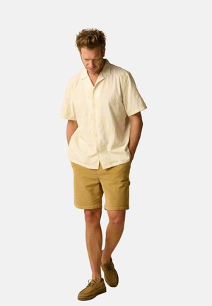 Homme portant une chemise crème à manches courtes avec des motifs floraux subtils, un short moutarde et des chaussures lacées beige, marchant les mains dans les poches.