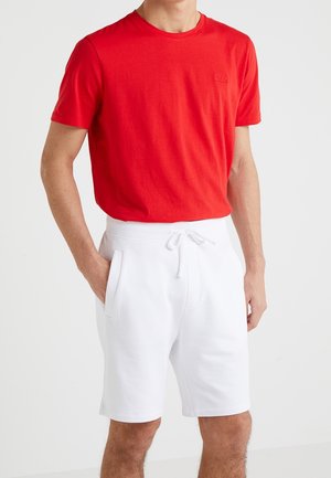 Homme portant un t-shirt rouge uni à manches courtes et un short blanc à cordon de serrage avec des poches, debout devant un fond uni.