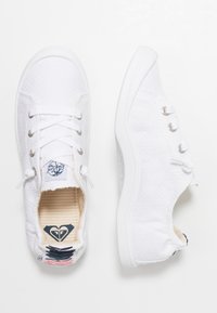 White canvas sneakers with a textured upper, round toe, and laces. Features silver eyelets and a patterned insole. Minimalist design.