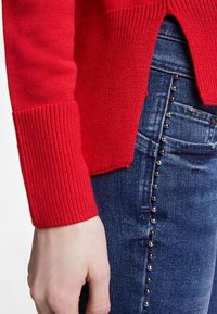 Roter Strickpullover mit gerippten Bündchen, kombiniert mit dunkelblauen Jeans aus Denim, die silberne Nietenakzente entlang der Seitennaht aufweisen.