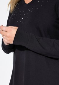 Musta pitkähihainen paita V-pääntiellä, jossa on pieniä metallinappeja yläosassa ja sileä, pehmeä rakenne.