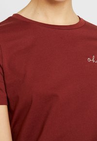 Burgunderfarvet T-shirt med rund halsudskæring, korte ærmer og en broderet detalje med teksten "oh" i hvid på øverste venstre side. Glat tekstur.