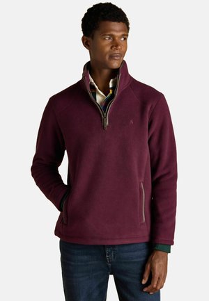 Mann trägt einen weinroten Fleece-Pullover mit Viertelreißverschluss, hat die Hände in den Taschen und trägt dazu eine dunkelblaue Jeans. Er schaut leicht nach rechts.