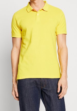 Hombre con una camiseta polo amarilla brillante de manga corta combinada con vaqueros azul oscuro, de pie frente a un fondo claro y liso.