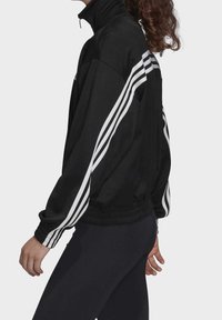 Black jacket with a high collar, featuring three white stripes along the sleeves. Soft fabric with a relaxed fit and elastic cuffs.