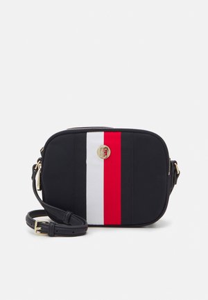 Small black crossbody bag with adjustable strap, vertical white and red stripes, and round gold logo emblem on front.