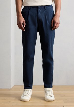 Man wearing navy blue slim-fit trousers and white sneakers standing on wooden floor against plain wall.