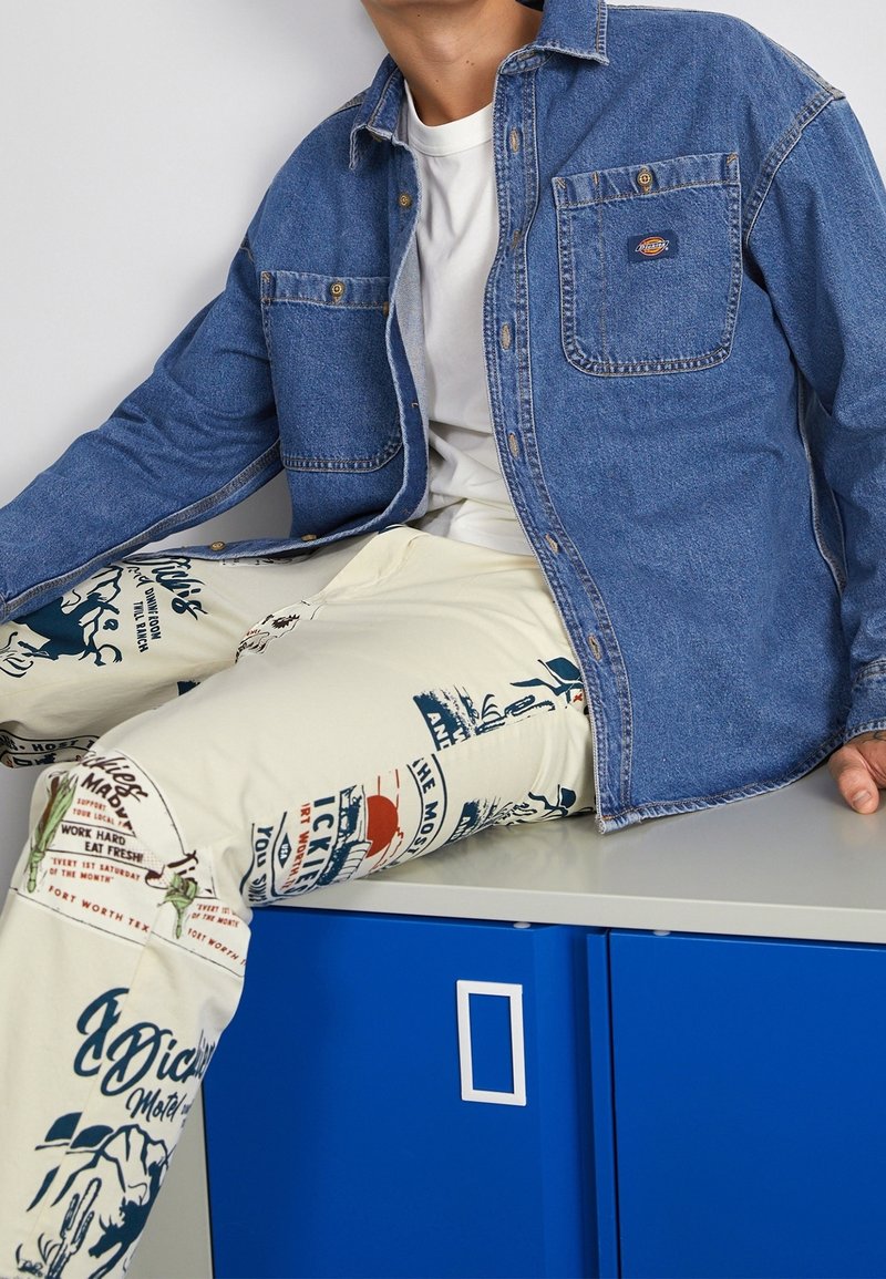 La camicia in denim con bottoni presenta una tasca sul petto e cuciture grigie. I pantaloni crema sono decorati con stampe grafiche blu e rosse e testo.