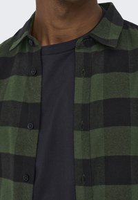 Chemise en flanelle à carreaux verts et noirs avec un t-shirt uni foncé en dessous. Elle comporte des boutons et un col. Texture douce avec un design décontracté.