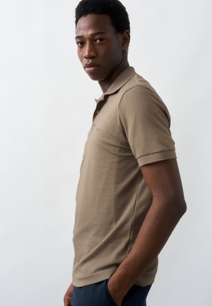 Νέος άνδρας με σκουρόχρωμο δέρμα και κοντά μαλλιά, φορώντας μπεζ πόλο πουκάμισο και σκούρο παντελόνι, στέκεται πλευρικά με το ένα χέρι στην τσέπη.