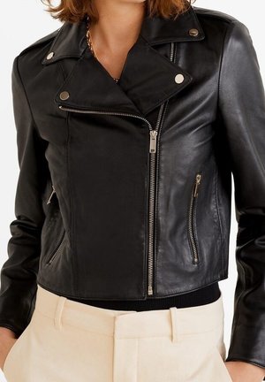 Mujer con una chaqueta de cuero negra ajustada de estilo motorista, con cremalleras plateadas y pantalones beige de cintura alta, cropé en la cintura.