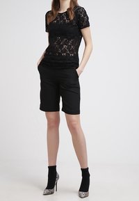Top corto in pizzo nero a maniche corte con motivo floreale, abbinato a pantaloni corti neri su misura. Il modello indossa calzini neri alla caviglia e tacchi con stampa argento.