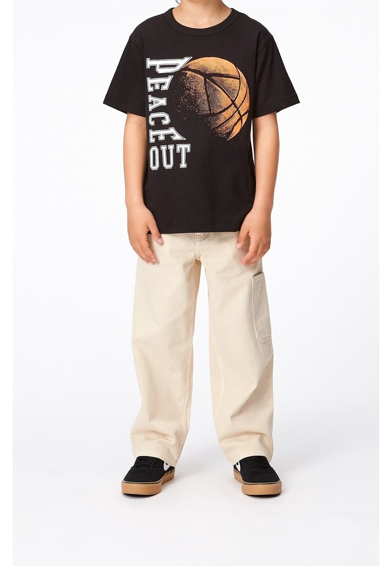 Enfant portant un T-shirt noir avec le texte "Peace Out" et un graphisme de basket-ball, un pantalon cargo beige et des baskets noires avec des semelles en gomme.