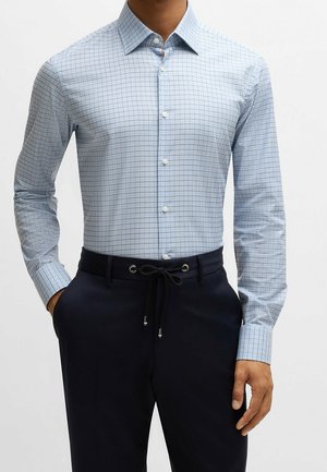 Man draagt een lichtblauw geruit overhemd met lange mouwen, ingestopt in donkerblauwe broek met trekkoord.