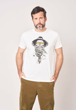 Man met baard die een wit T-shirt draagt met een afbeelding van een gebaarde schedel met zonnebril en hoed, gecombineerd met een bruine broek, staand met handen in de zakken.