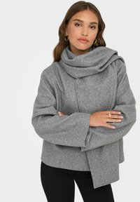 Grå ullig omslagskofta med en stor, draperad krage, vida ärmar och en boxig passform, kombinerad med svarta byxor och en ring på ena handen.