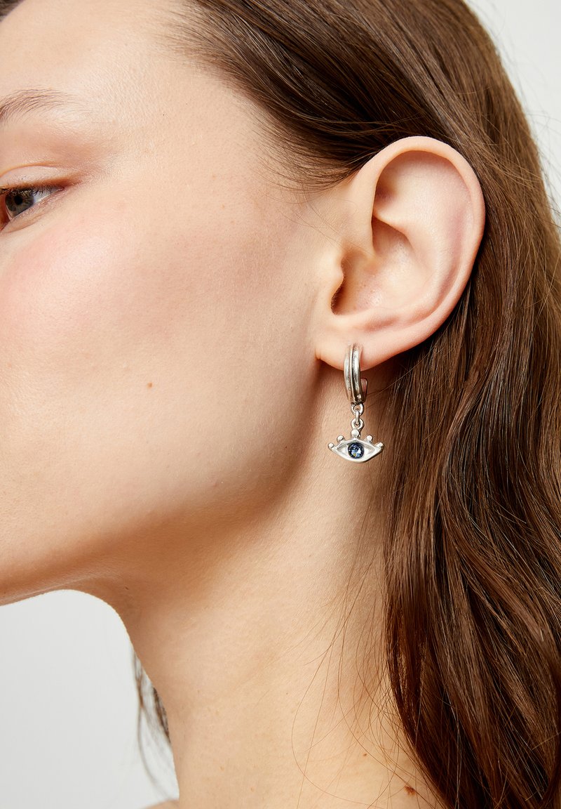 Pendiente de aro plateado con un colgante en forma de ojo que presenta una piedra azul. El diseño incluye pequeños detalles elevados alrededor del ojo.
