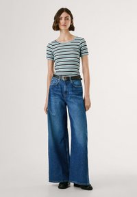 Woman wearing a light blue striped t-shirt, wide-leg blue jeans with a black belt, and black shoes, standing against a white background.
