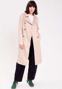 Cappotto trench in suede beige doppiopetto con bottoni marroni lucidi, abbinato a jeans scuri e sneakers verdi. Maniche lunghe e design a fronte aperta.