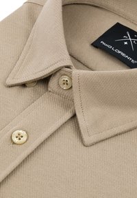 Beżowa koszulka polo o tkanej fakturze, z dwoma guzikami i kontrastującą czarną metką z logo. Kołnierz ma klasyczny design.