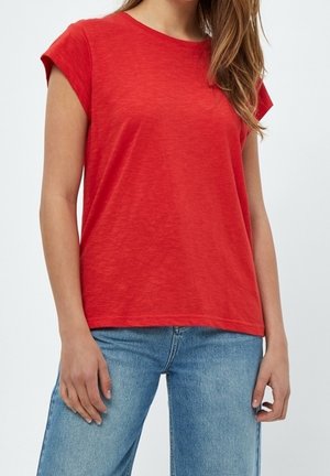 Kobieta w prostym czerwonym t-shircie z krótkim rękawem i jasnoniebieskich dżinsach stojąca na białym tle.