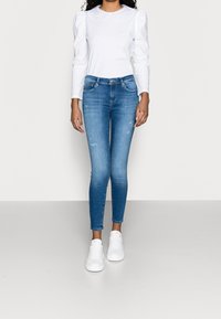 Mujer vestida con una blusa blanca de manga larga, jeans ajustados azules y zapatillas blancas, de pie contra un fondo gris claro.