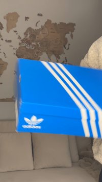 Scatola di scarpe Adidas blu con strisce bianche tenuta davanti a un divano beige e a una mappa del mondo marrone su una parete grigia.